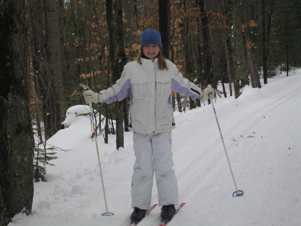 Young girl wearing white ski suite and blue hat on skis and holding poles on snowy cross country ski trail through the woods at Chateau Montebello.