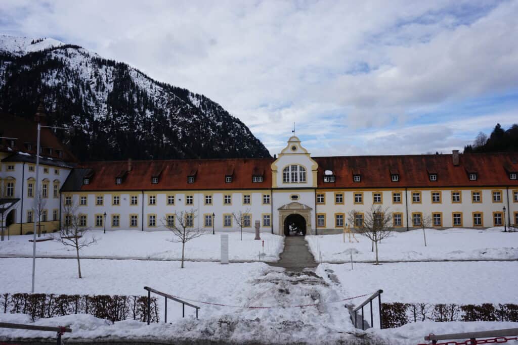 Building on grounds of Ettal Monastery with snow on ground and mountain in background.