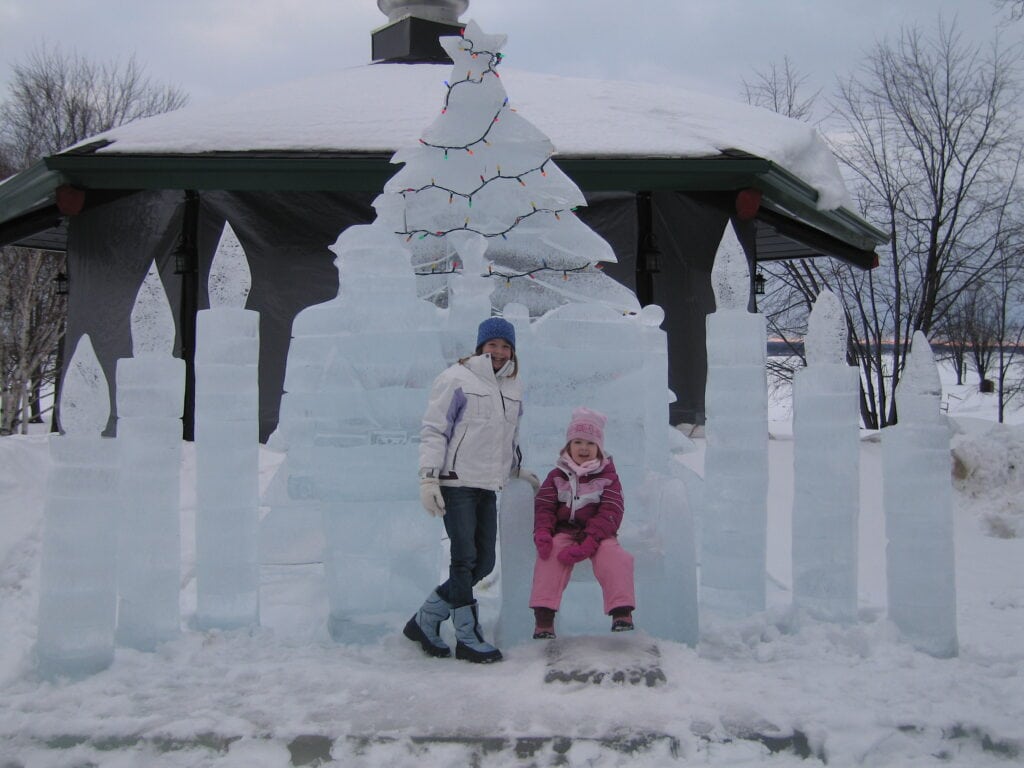 Two girls standing in front of ice sculpture at Chateau Montebello in Quebec. Older girl wearing jeans, white ski jacket and blue hat is standing. Younger girl wearing light pink snow pants, dark pink jacket and light pink hat is sitting on chair carved from ice.