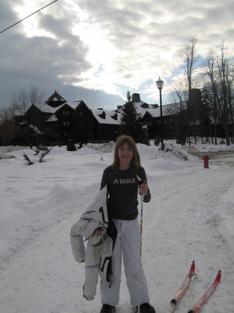 Young girl wearing white ski pants and dark shirting holding white ski jacket and ski pole with skis on the ground standing on snowing trail in front of Chateau Montebello.