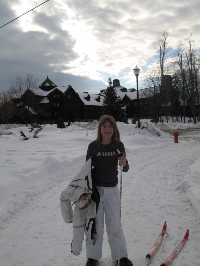Young girl in dark top and white snow pants hlding ski poles and white jacket with skiis on ground beside her and Chateau Montebello in background.