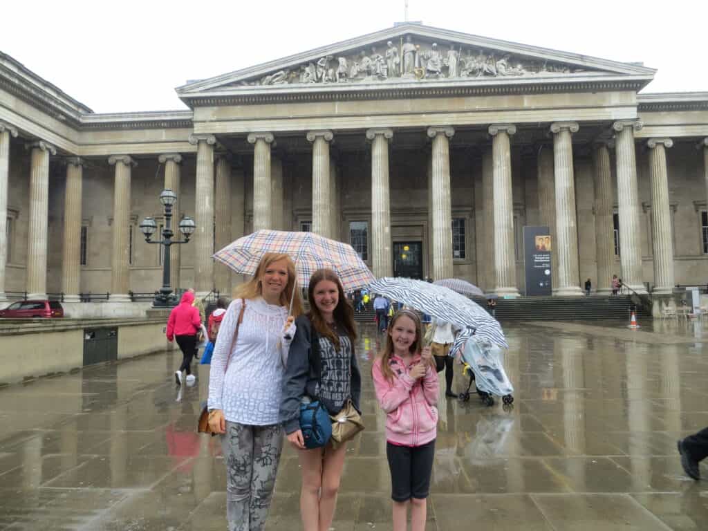 Two teenage girls and a younger girl holding umbrellas in rain in front of the British Museum in London.