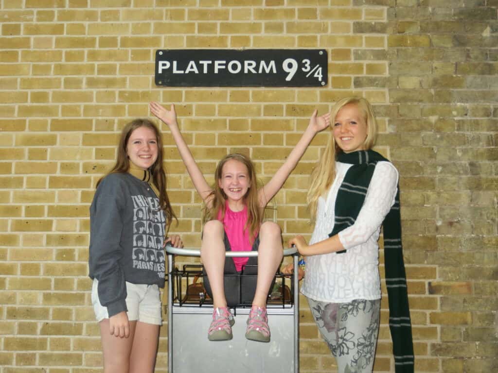 Two teenage girls and a young girl sitting in a luggage cart between them in front of a brick wall and a sign reading Platform 9 3/4.