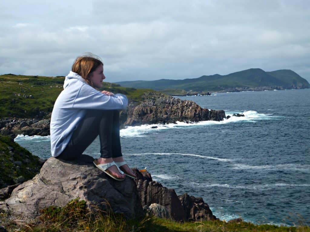 Teen girl wearing jeans, grey sweatshirt and white sandals sitting on rocks looking out at ocean at Ferryland Lighthouse, Newfoundland.