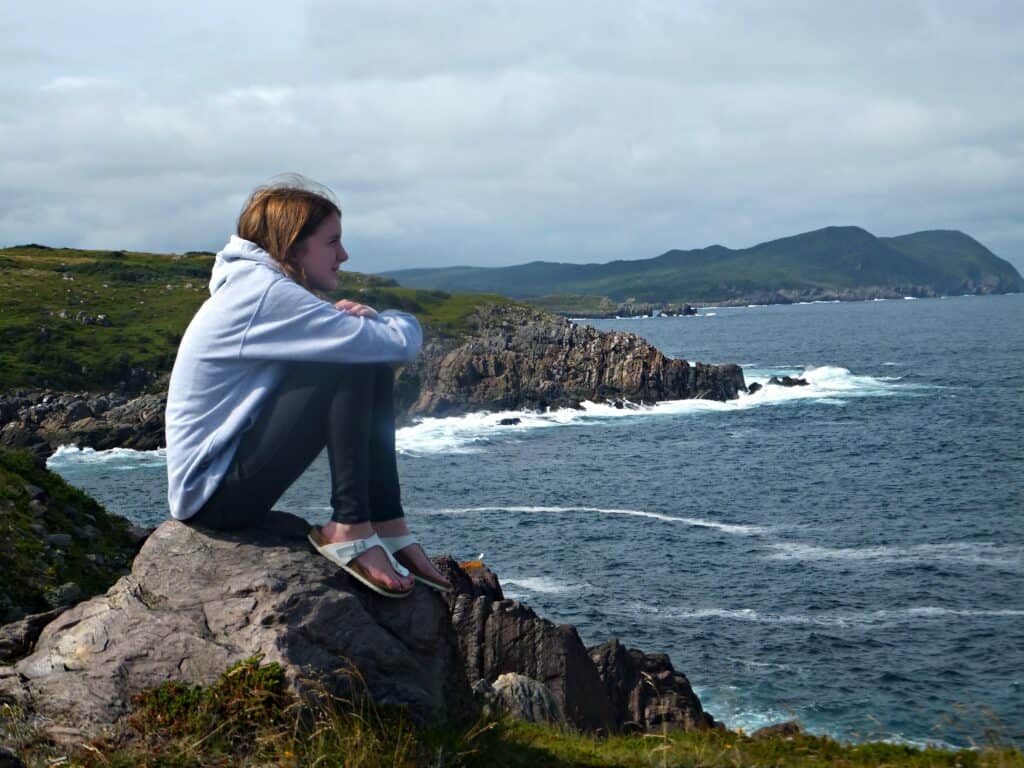 Teen girl wearing dark jeans, grey sweatshirt and white sandals sits on rocks hugging knees looking at ocean at Ferryland, Newfoundland.