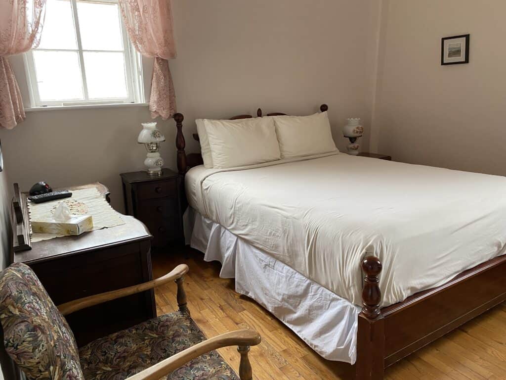 Tower Room at West Point Lighthouse Inn - old-fashioned room with hardwood floors, bed with sheets and pillows, dresser, chair and night table with glass lamp, pink lace curtains.