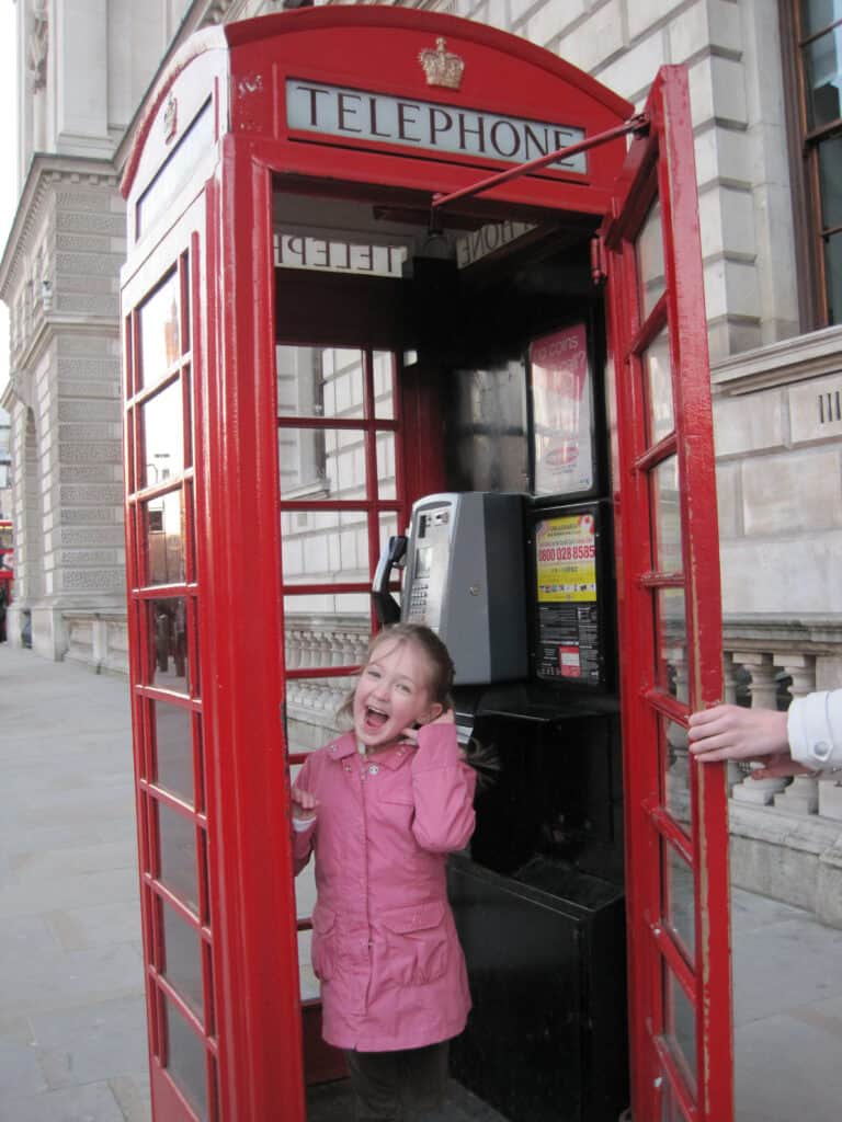 Young girl in pink coat pretending to talk on phone in red telephone booth in London, England.