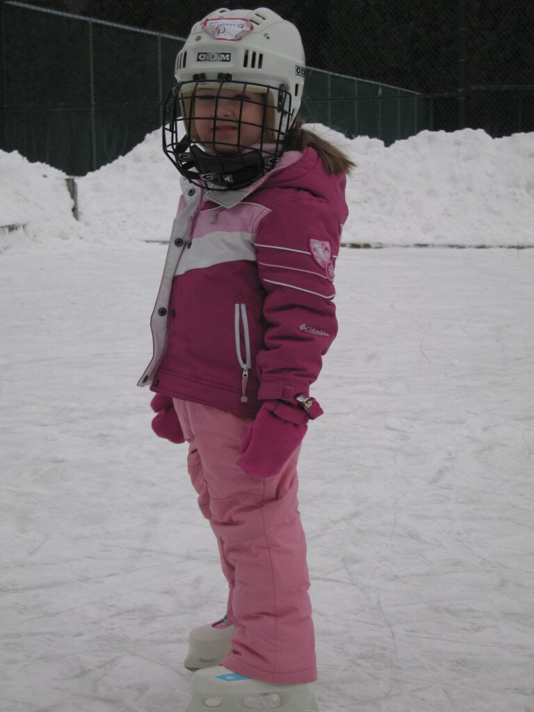 Young girl in pink ski suite wearing white hockey helmet with black face guard and skates on outdoor skating rink at Chateau Montebello.