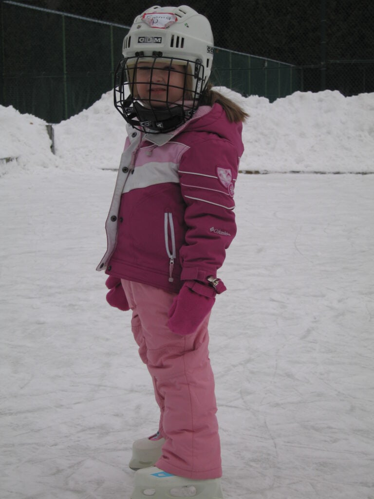 young girl in pink snow suit, white skates and hockey helmet on outdoor skating rink - Chateau Montebello.