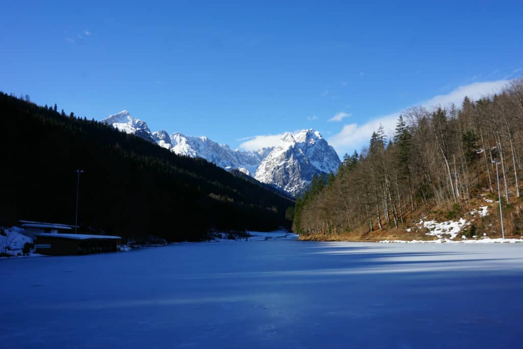 Alpine lake in Garmisch, Germany - ice and snow covered lake with bare trees and snow-topped mountain peak in background.