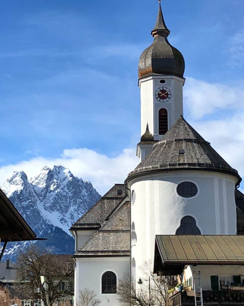 White church with dark roof and domed steeple and clock in Garmisch-Partenkirchen with snow-capped mountain peak in background and blue sky with a few clouds.