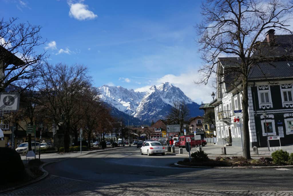 Street scene in Garmisch-Partenkirchen with buildings, cars, bare trees and snow-capped mountain peak in background.