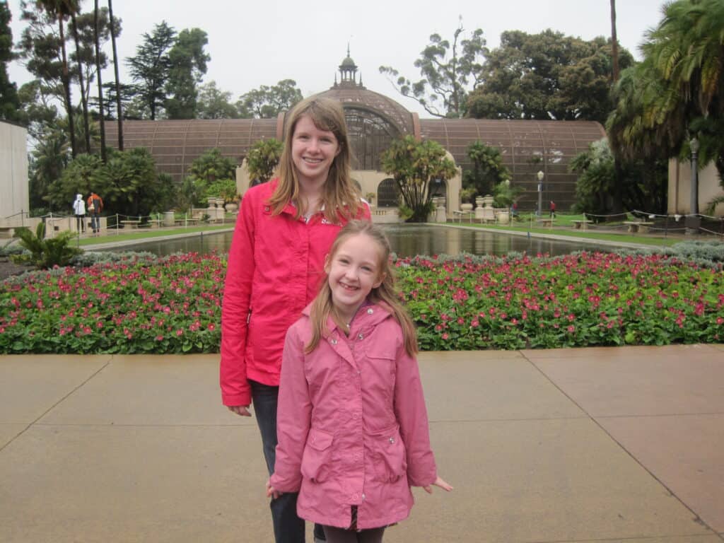 Two young girls standing in Balboa Park, San Diego on a rainy day in front of pond and flower garden.