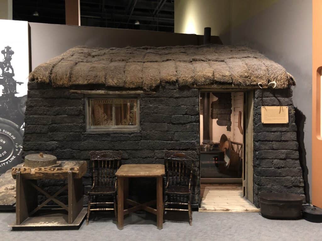 Dark cabin with sod roof, wooden table and chairs outside entrance on display at the Manitoba Museum.