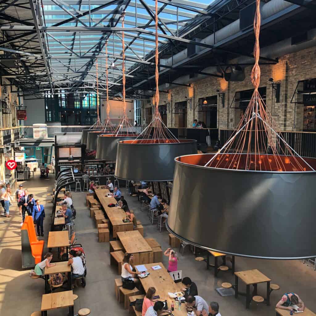 View of the Food Hall at The Forks Market from the second level - large black drum lights hanging from ceiling and people sitting at light coloured wooden tables on the first level.
