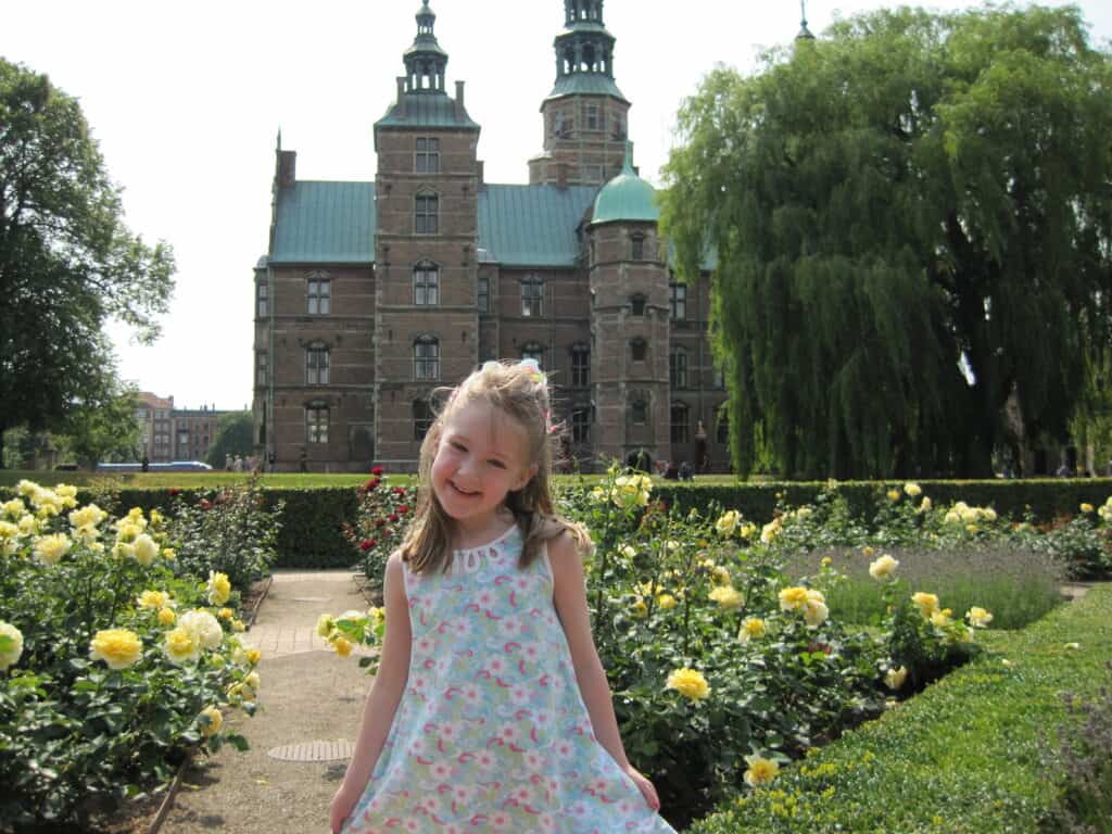 Young girl in blue and pink flowered dress standing in gardens with yellow roses and Rosenborg Castle in Copenhagen in the background.