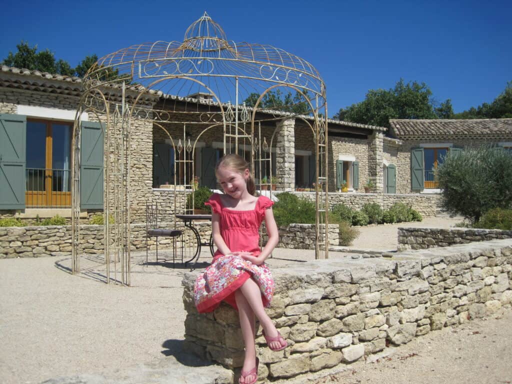 Young girl with ponytail wearing coral top and flowered skirt sitting on stone wall in front of stone villa with sage green shutters in Gordes, France.