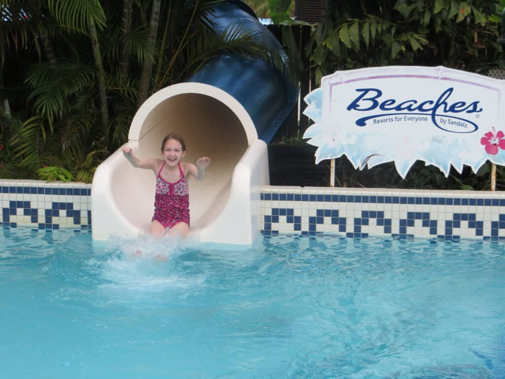 Young girl in dark pink flowered bathing suit entering pool from waterside next to Beaches Resort sign in Negril, Jamaica.
