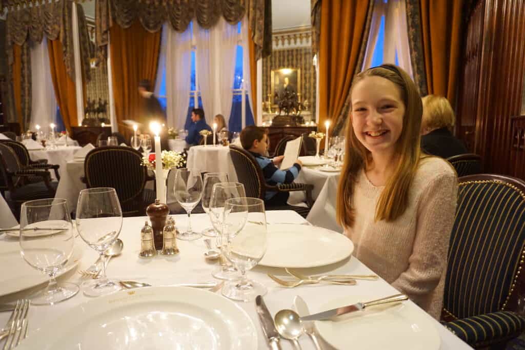 Girl in light coloured sweater sitting on dark pin-striped chair at a table set with white tablecloth, white plates and glassware and guests at more tables in background.