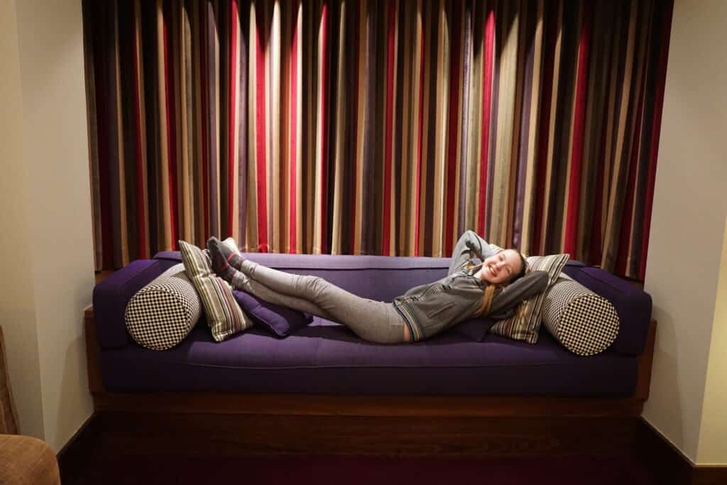Girl laying on eggplant coloured sofa with bolsters and cushions in front of striped multi-coloured closed curtains.
