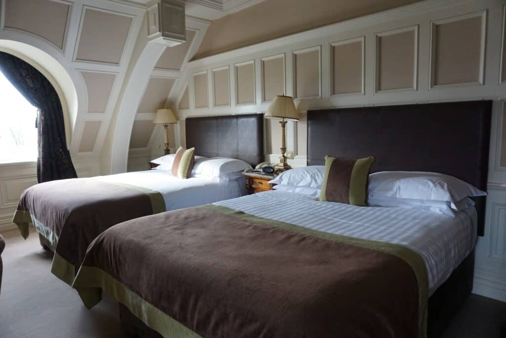 O'Connell Suite at Killarney Park Hotel - beige and cream panelled woodwork, curved window with dark curtain, two double beds with dark headboards, white sheets and dark brown and green blanket over foot of bed with two bedside tables with lamps.