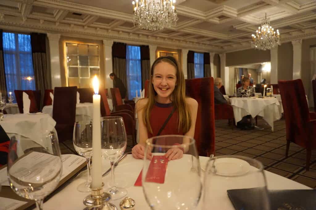 Girl in burgundy dress sitting on burgundy velvet highback chair at round table with white tablecloth and glassware in The Park Restaurant at Killarney Park Hotel with additional tables and diners in background.