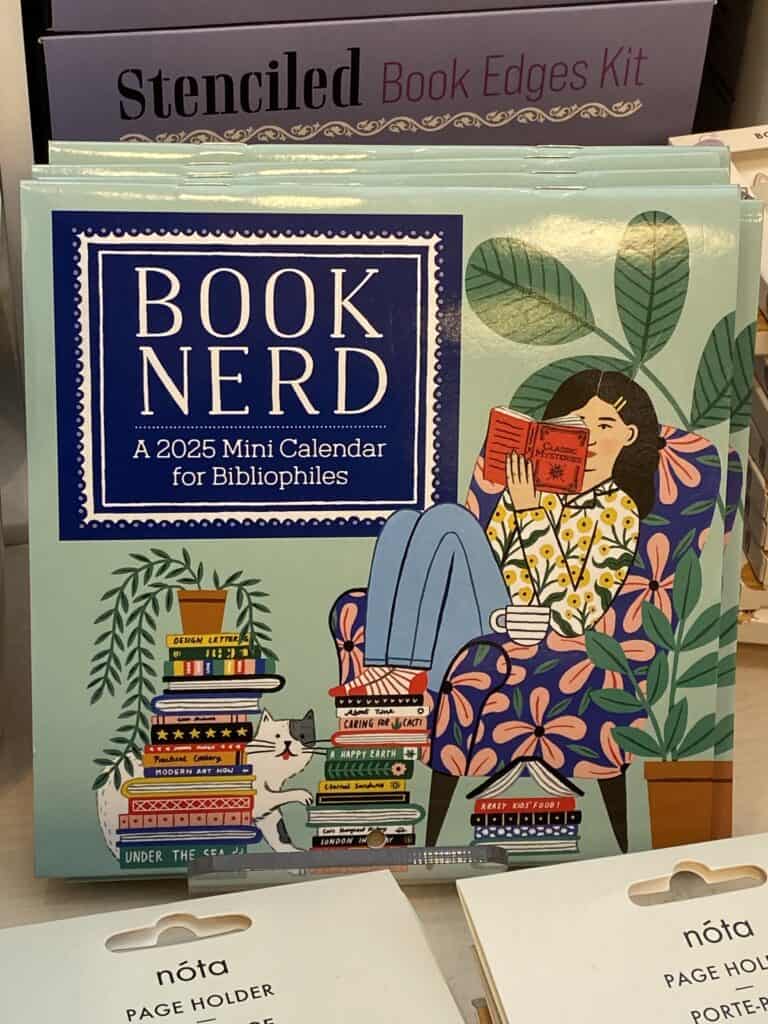 Cover of Book Nerd - A 2025 Mini Calendar for Bibliophiles on display - illustration of a woman sitting in a chair reading with a stack of books on floor.
