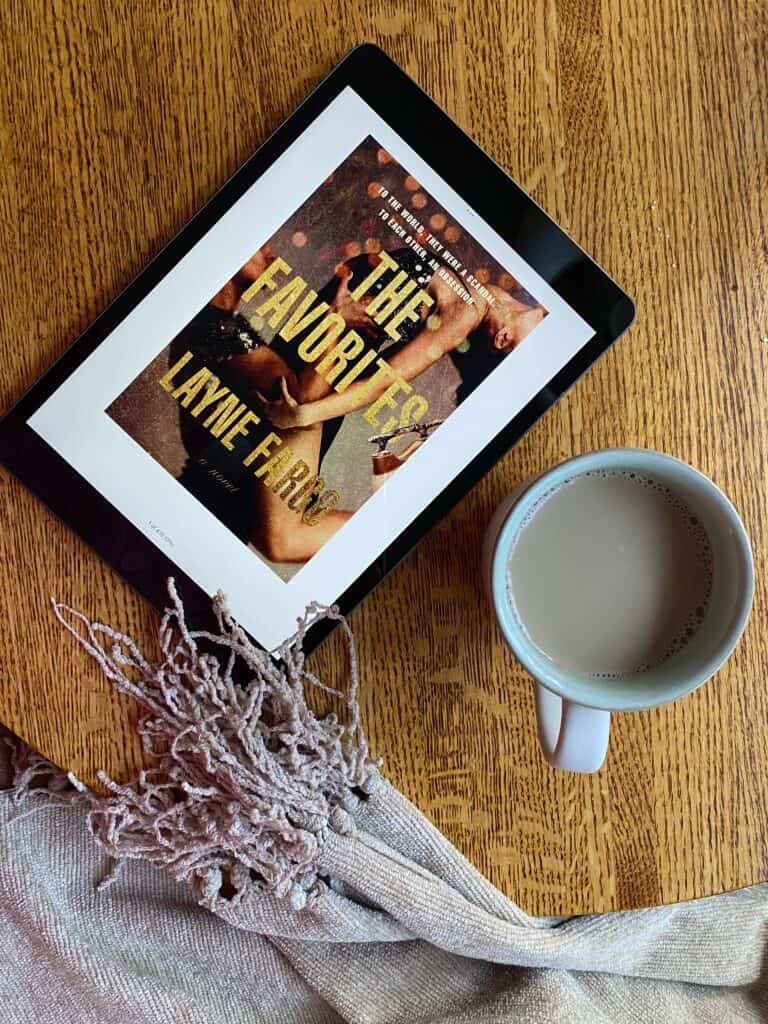 A blanket, cup of coffee and iPad displaying book cover for The Favorites by Layne Fargo sitting on a wood table.