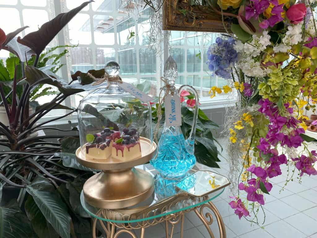 Small glass top table with glass bottle containing blue liquid and tag reading 'Drink Me' and small covered cake stand with squares of dessert with tag reading "Eat Me' surrounded by colourful flowers and greenery.