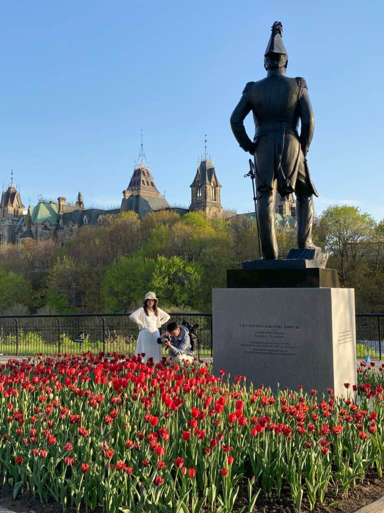 Statue of Colonel By in Major's Hill Park surrounded by bed of red tulips and Parliament in background. Man with camera kneels taking photo and woman in white dress, sweater and hat watches.