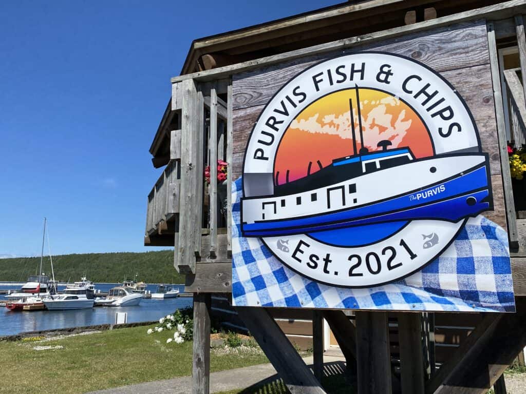 Sign for Purvis Fish & Chips on grey wooden building with boats in Gore Bay Marina in background.