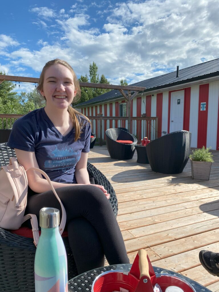 Young woman sitting in wicker chair on wooden deck with red and white stripe building in background at Campbell's Drive-in, Gore Bay, Ontario.