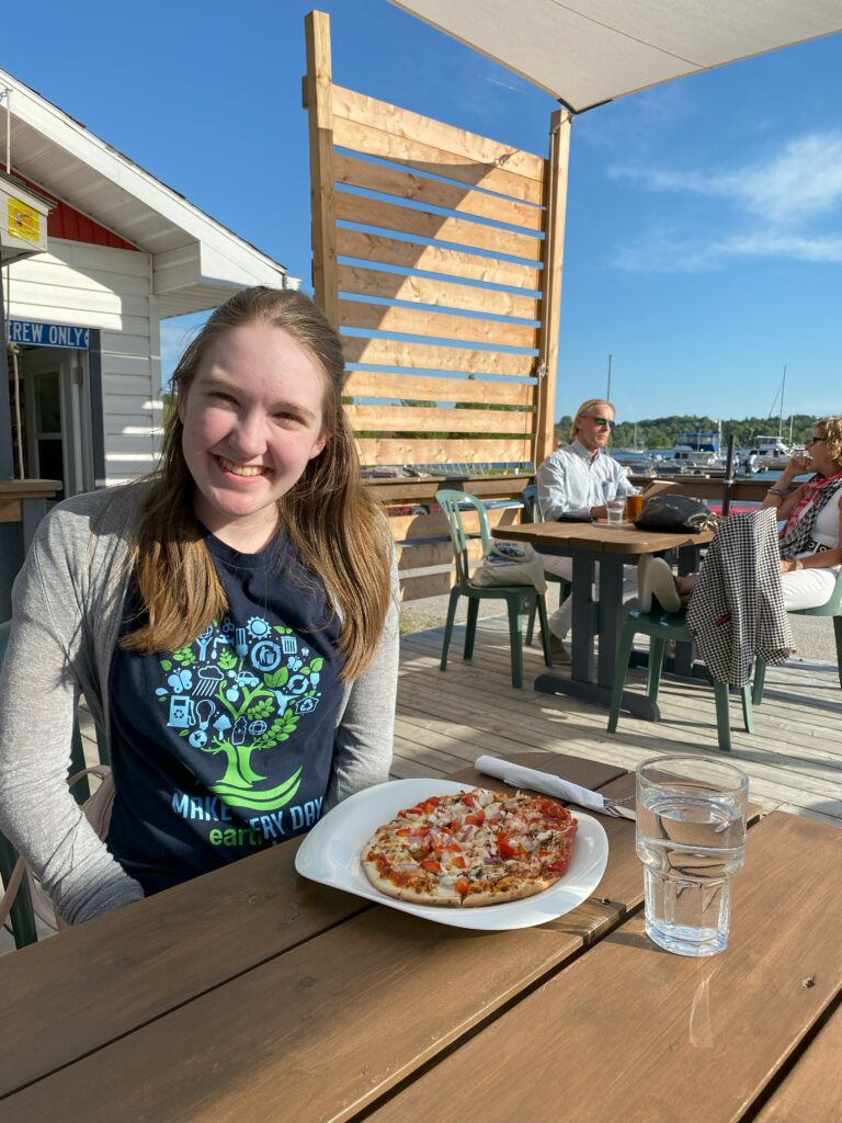 Young woman sitting on patio at Buoy's Restaurant in Gore Bay with small pizza on table in front of her and in the background a couple at another table and boats in the marina.