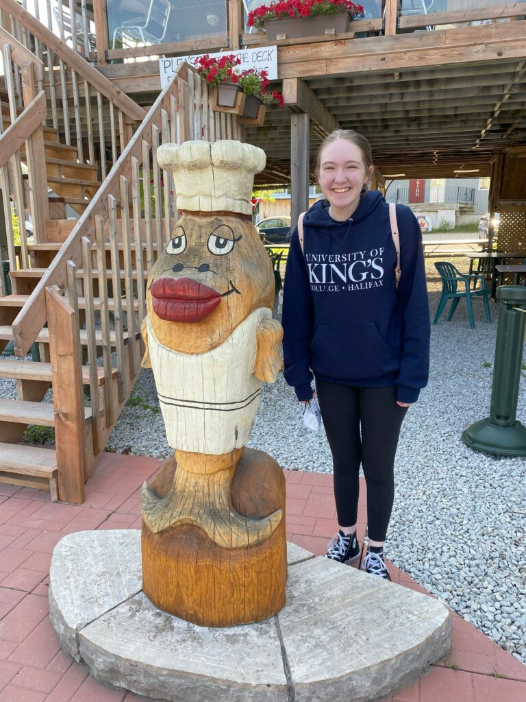 Young woman standing beside comical wooden sculpture of a fish outside The Codmothers in Gore Bay, Ontario.