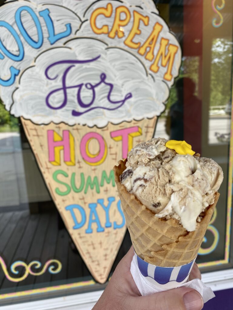 Hand holding ice cream cone in front of multi-coloured sign painted on door of ice cream cone reading 'Cool Cream for Hot Summer Days"