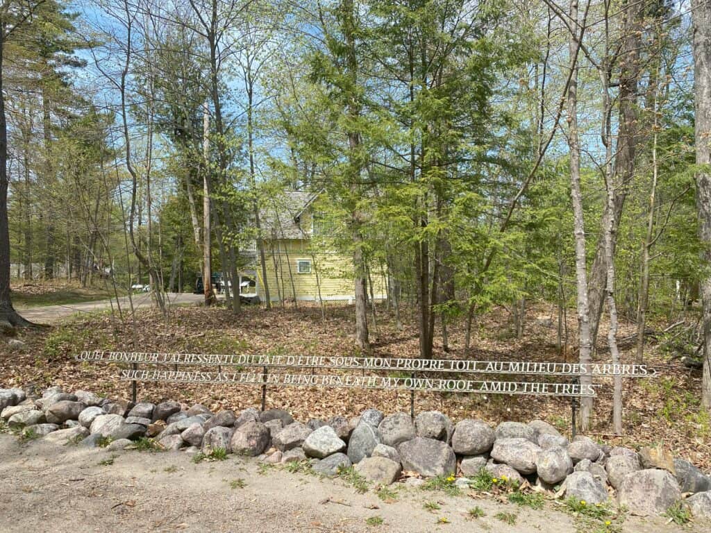 Clear plastic panel with etched quote in white mounted alongside dirt path lined with rocks in wooded area with building visible through trees in background.