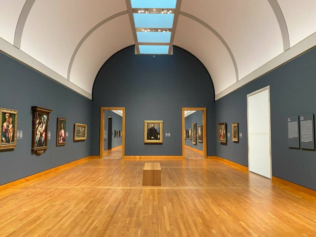 Gallery of paintings on display at the National Gallery of Canada - room has light hardwood floors, dark grey walls and curved white and grey ceiling.