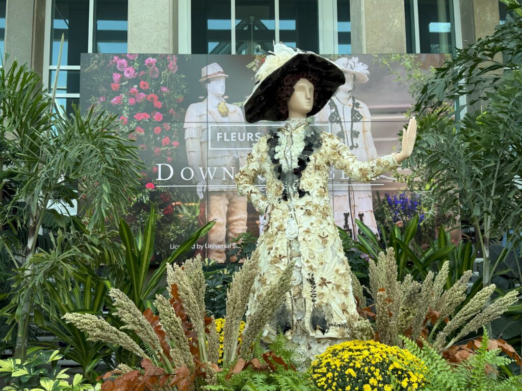 Flower mannequin in white gown with black color and large black and white hat surrounded by greenery in front of Fleurs de Villes: Downton Abbey background at entrance to the RBG.