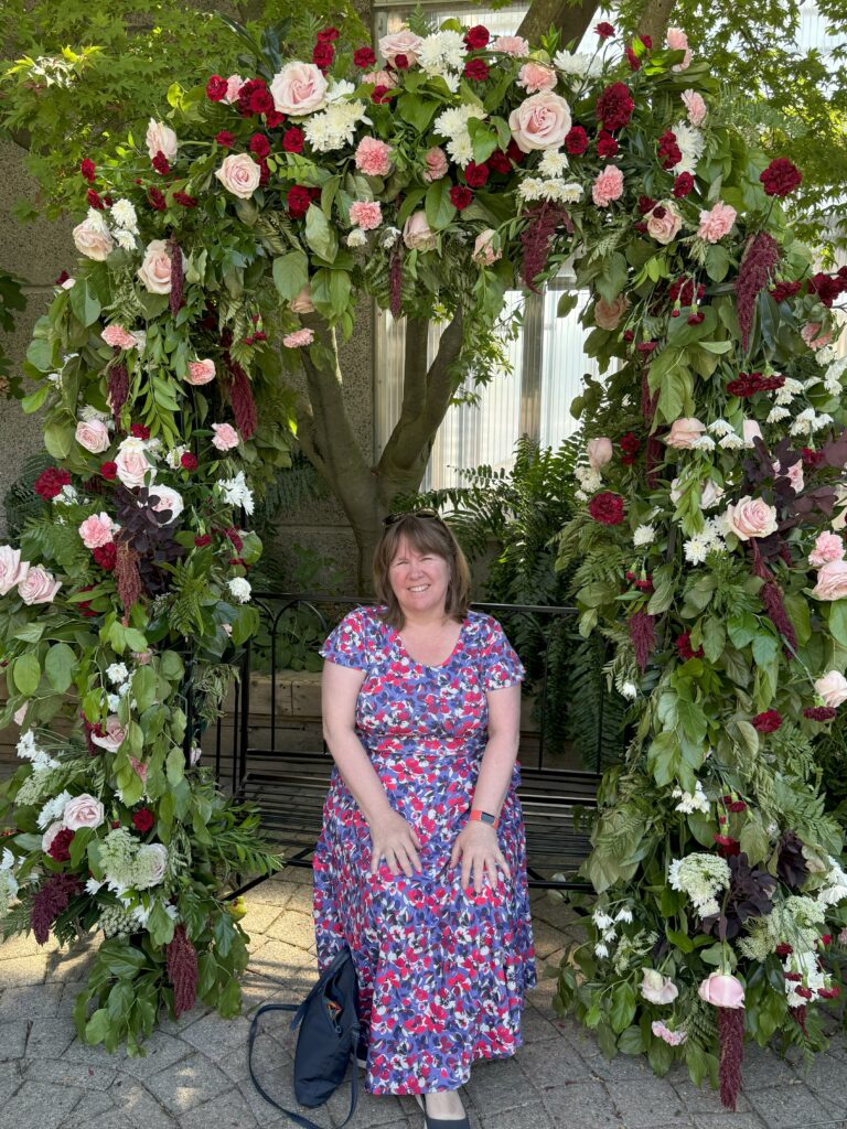 woman in blue, red and purple flowered dress sitting on bench surrounded by arch of pink, red, and white roses.
