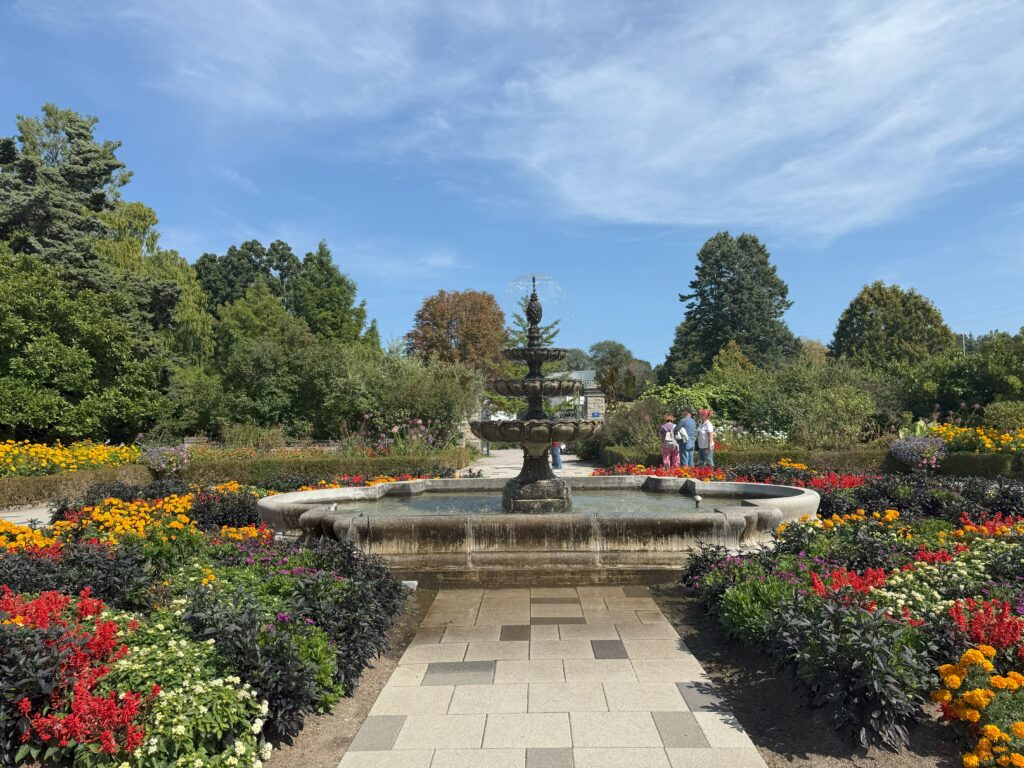 Three tier fountain in Hendrie Park at the Royal Botanical Gardens surrounded by beds of colourful flowers and trees set against bright blue sky with wispy white clouds.