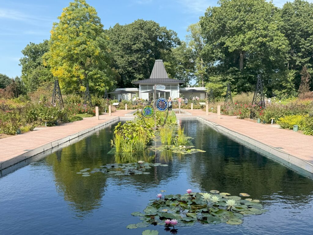 reflecting pool with lily pads, pink and white flowers and additional greenery surrounded by gardens with tea house in background.