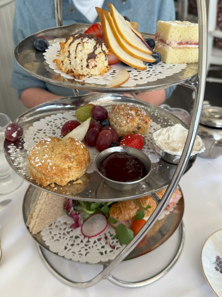 three tiered tray for afternoon tea with sandwiches on botom, two scones with clotted cream, jam and fruit in middle and small desserts and fruit on top.