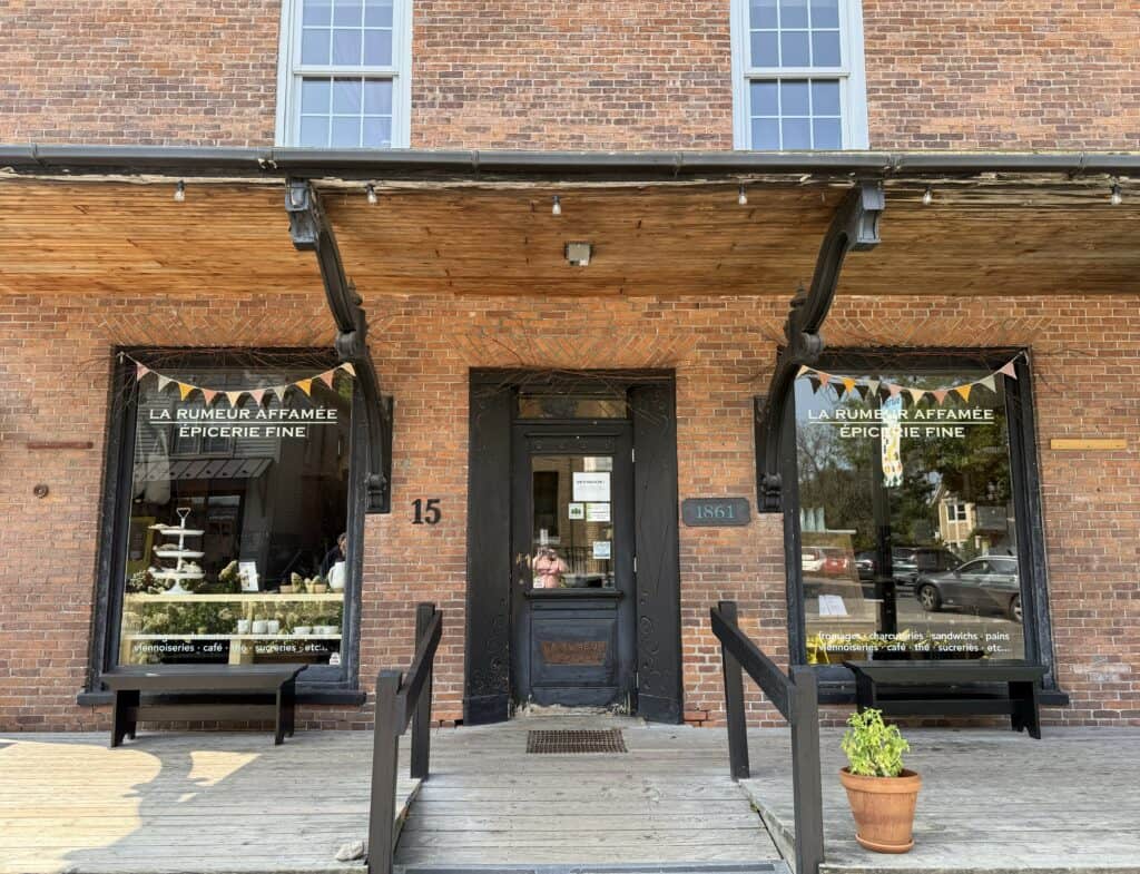 red brick building with two front windows with dark brown frames and dark front door - words on both windows read "La Rumeur Affamee Epicerie Fine".