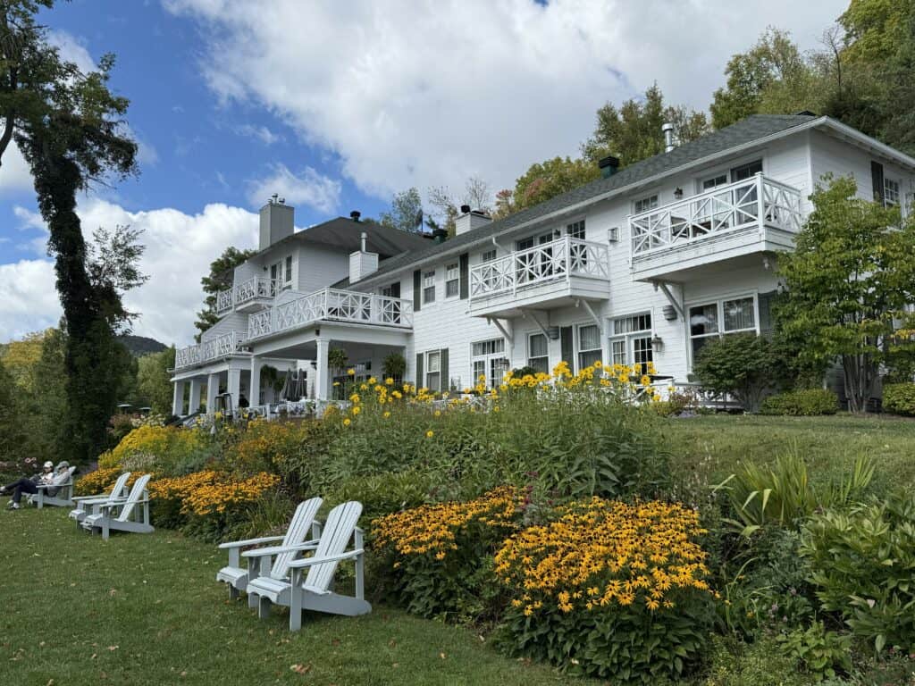 Exterior of Manoir Hovey a white building with dark roof, many windows, balconies and covered porch - with white adirondack chairs in front of flower beds on lawn - blue sky and white fluffy clouds.