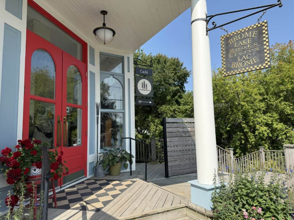 Entrance to Brome Lake Books. Light blue building with white trim and bright red door with large glass windows. Brome Lake Books sign hanging from white pole.