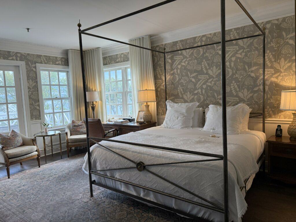 room at Manoir Hovey - black metal four poster bed with white linens and patterned wallpaper with seating area in front of large windows.