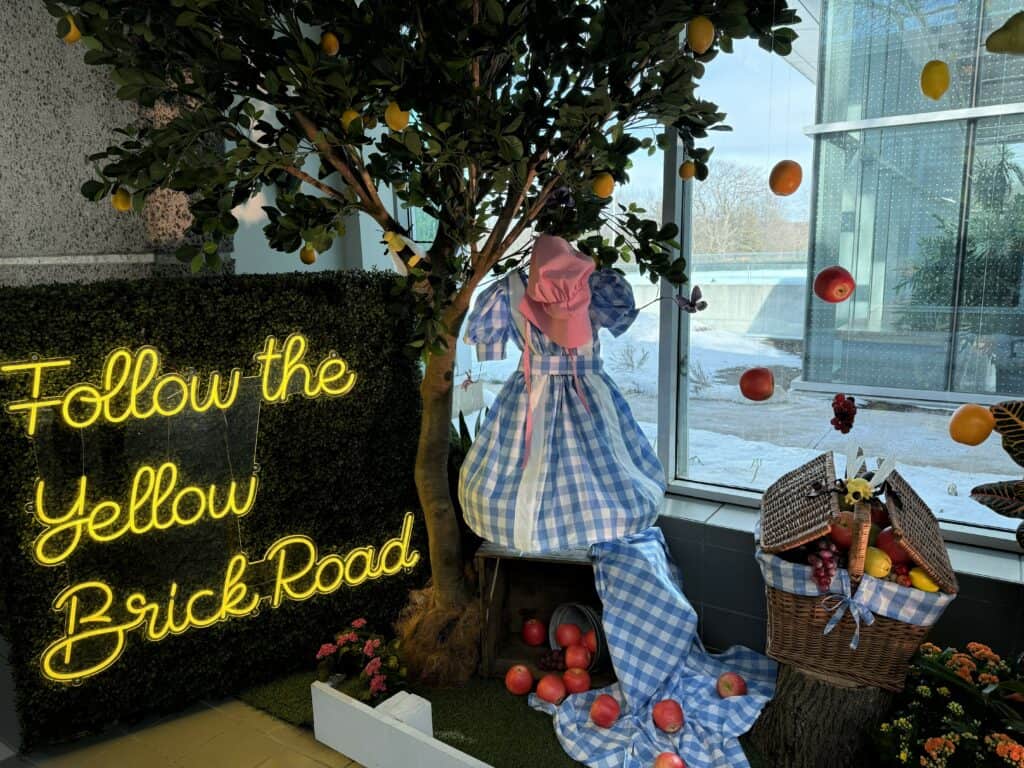 blue and white gingham dress, pink bonnet and picnic basket on display beside tree and sign reading "Follow the Yellow Brick Road" written in yellow cursive.