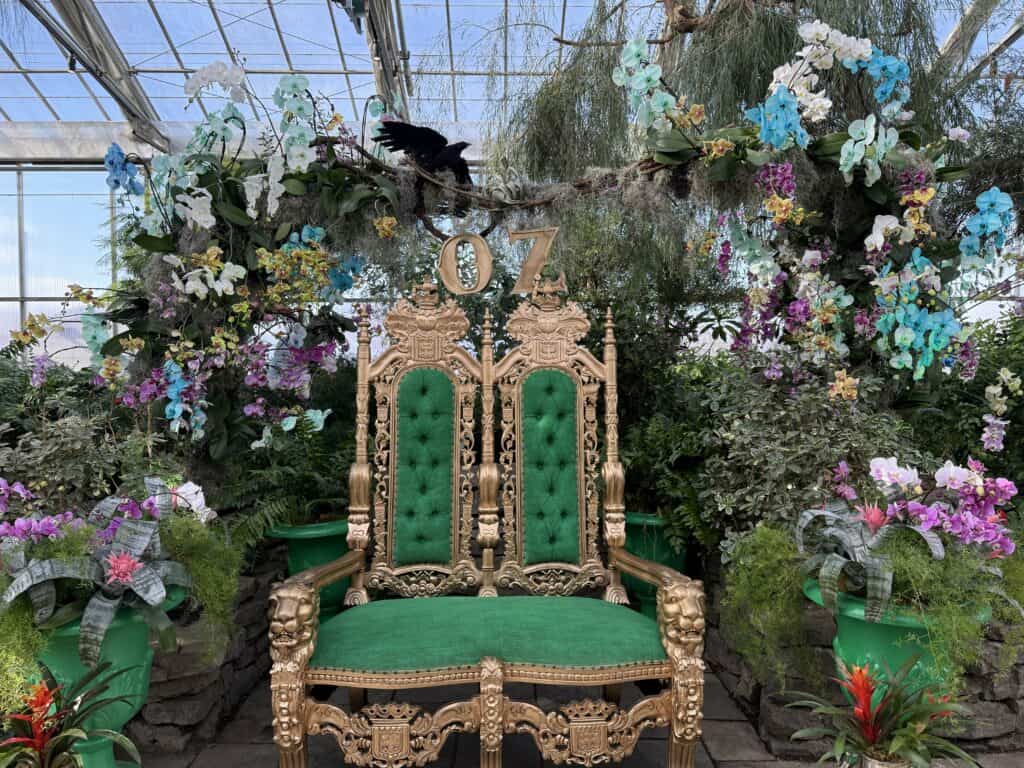 gold two person chair with green upholstery and the word "oz" at the top sitting amidst flowers and greenery.