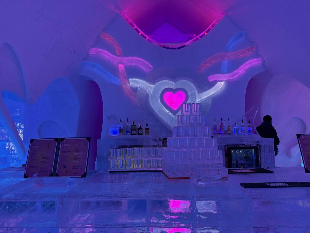 bar at quebec ice hotel with purple and pink lighting - glasses made of ice stacked on bar also made of ice - pink and purple heart on wall behind.