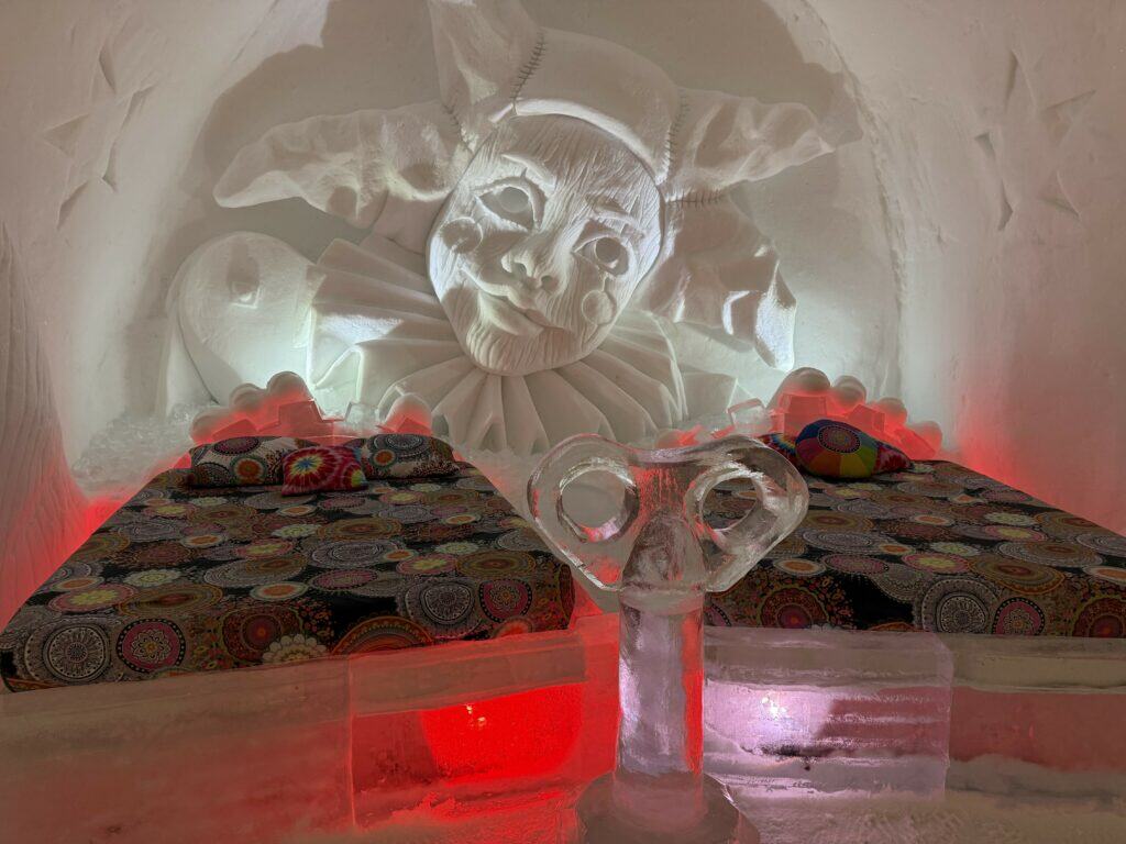 two double bed room at quebec ice hotel with colourful bedding and carving of clown in snow wall behind bed.